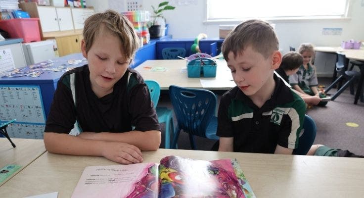 Two boys sitting at a desk in a classroom reading a book together.
